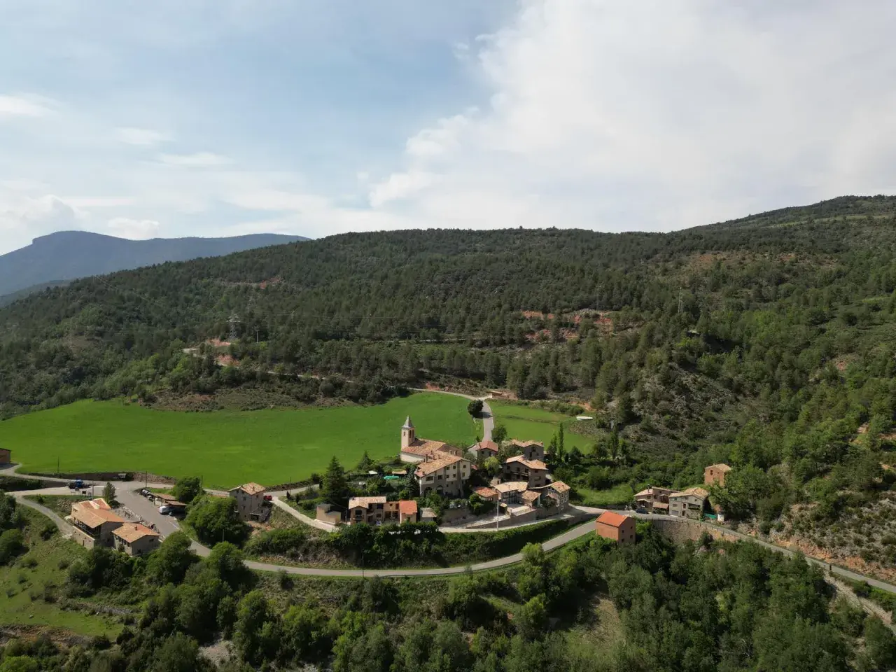Vista aérea del pintoresco pueblo de **Vall de la Vansa**, rodeado de verdes prados y frondosos bosques, con casas de piedra y una iglesia.