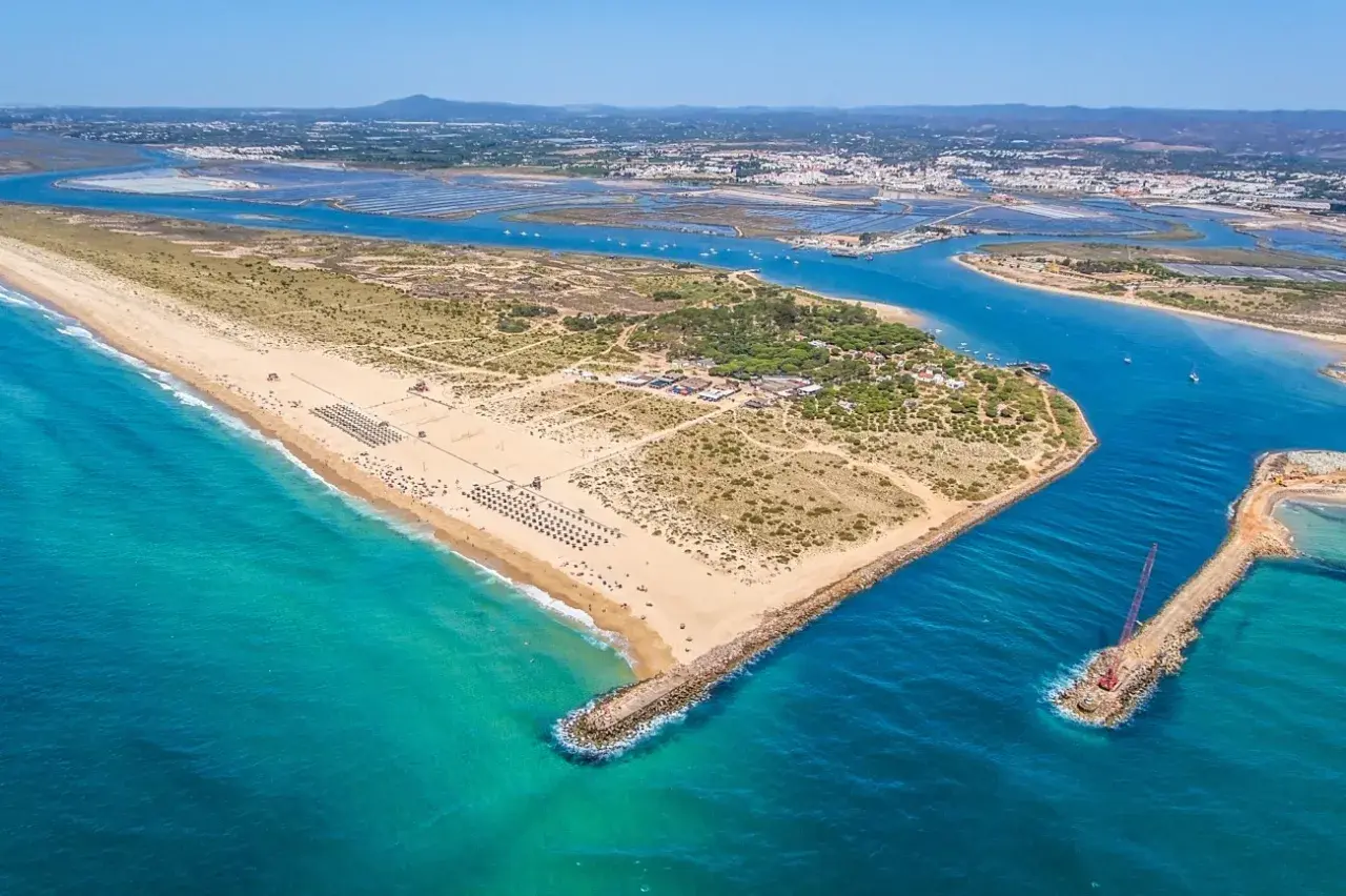 Vista aérea de la **isla de Tavira**, con su extensa playa de arena dorada, aguas turquesas y el canal que la separa de tierra firme, donde se aprecian salinas y la ciudad al fondo.