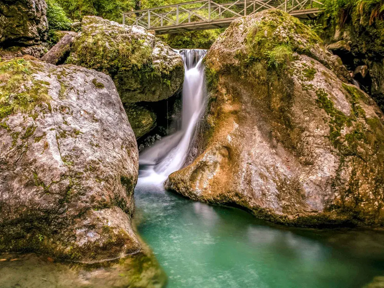 Cascada en las gargantas de Kakueta, con agua turquesa y rocas cubiertas de musgo. Un puente metálico cruza sobre la caída de agua.