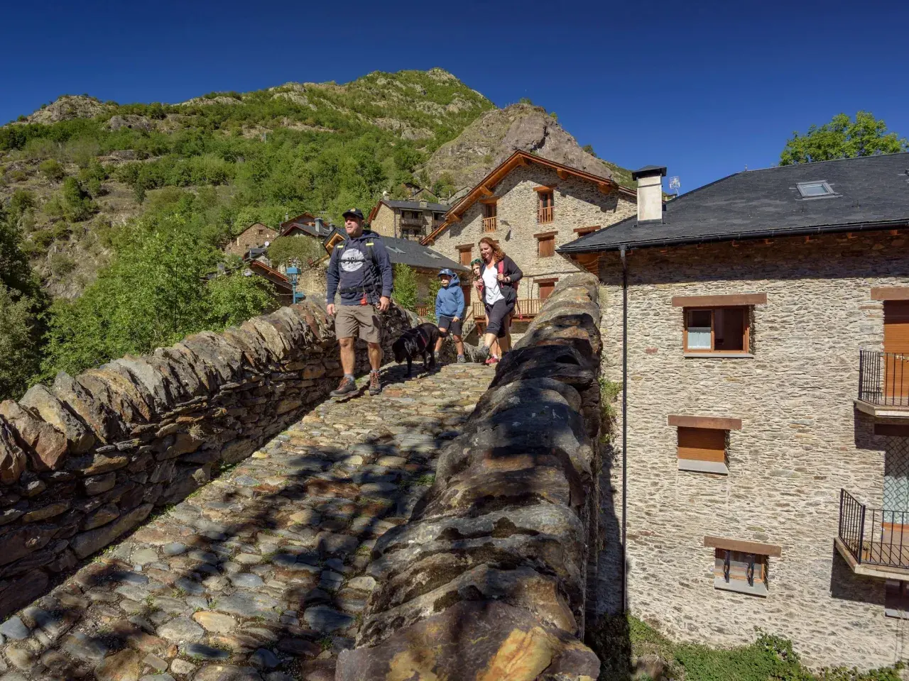 Familia pasea por un puente de piedra en un pueblo pirenaico, disfrutando de actividades al aire libre.