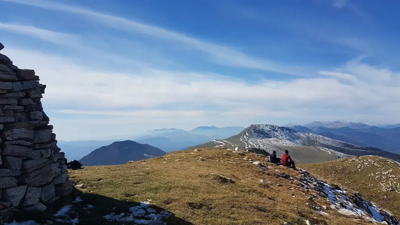 Dos personas contemplan el paisaje montañoso desde el Puig d'Estela, con un muro de piedra a la izquierda y el cielo azul con nubes.