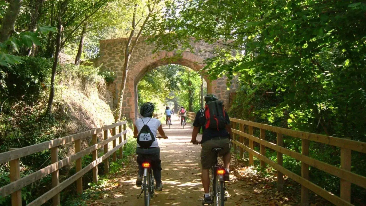 Ciclistas disfrutan de la vía verde Olot Girona, pedaleando bajo un antiguo arco de piedra rodeado de exuberante vegetación.
