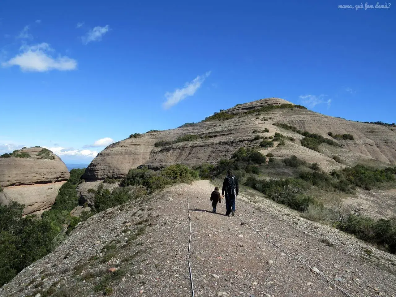 Un padre y su hijo caminan por un sendero rocoso hacia la cima del Montcau, bajo un cielo azul con nubes dispersas.