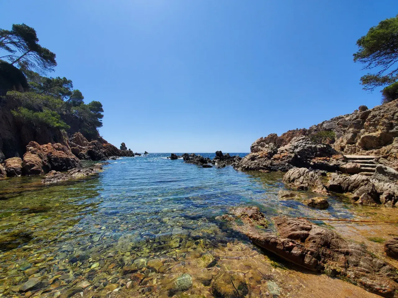 Aguas cristalinas y rocas rojizas en una cala idílica, un tesoro entre las mejores calas de la Costa Brava.