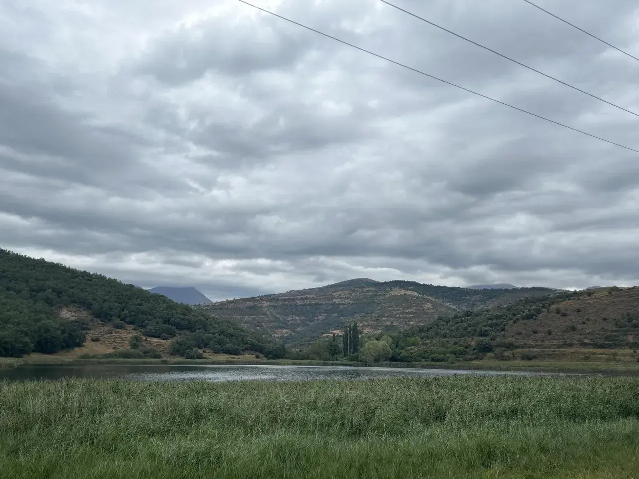 Paisaje sereno con un lago rodeado de colinas verdes y secas, bajo un cielo nublado. Las montañas a lo lejos parecen la geganta adormida.