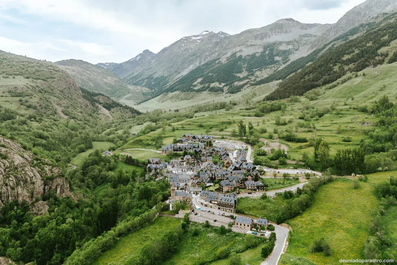 Pueblo de casas de piedra en un valle verde del Val d'Aran, rodeado de montañas cubiertas de vegetación y algunas cumbres nevadas.