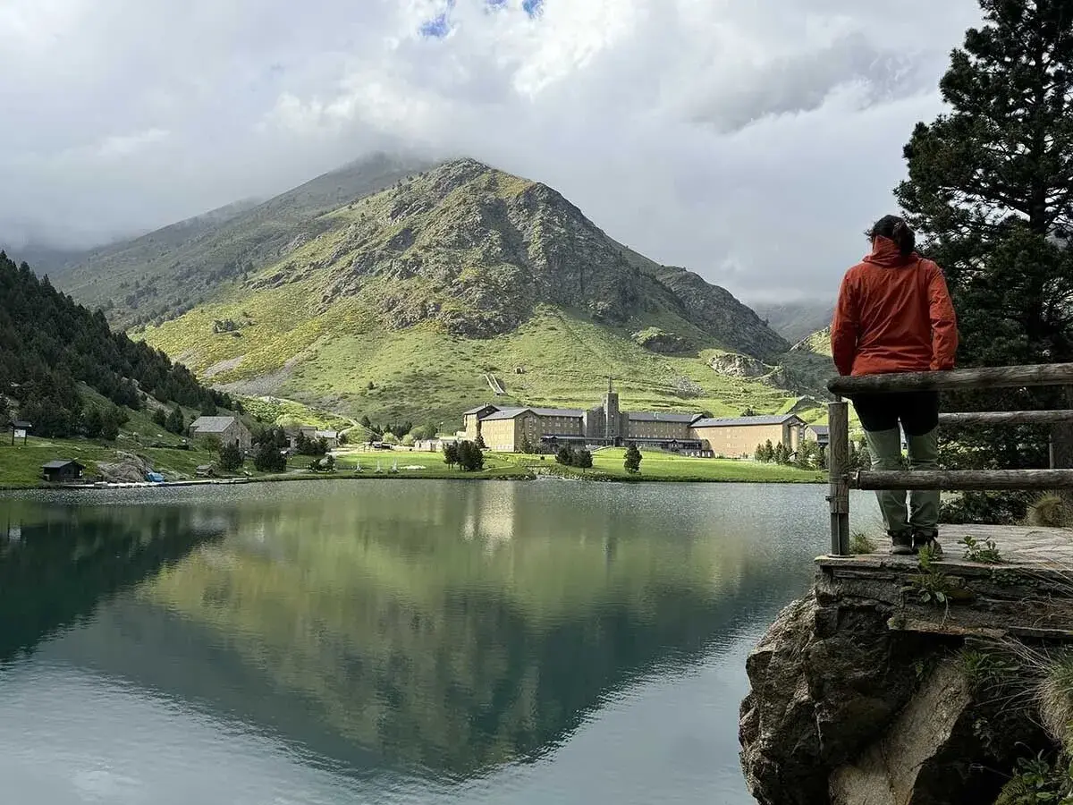 Persona contempla el sereno lago en el valle de Nuria, con imponentes montañas verdes y un complejo de edificios al fondo.