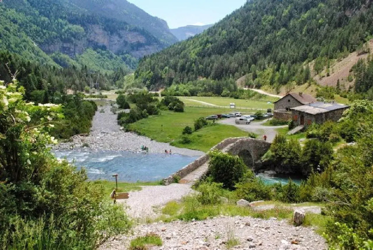 Un río de aguas turquesas fluye por un valle verde, cruzado por un puente de piedra. La ruta Bujaruelo invita a disfrutar de este paisaje montañoso.
