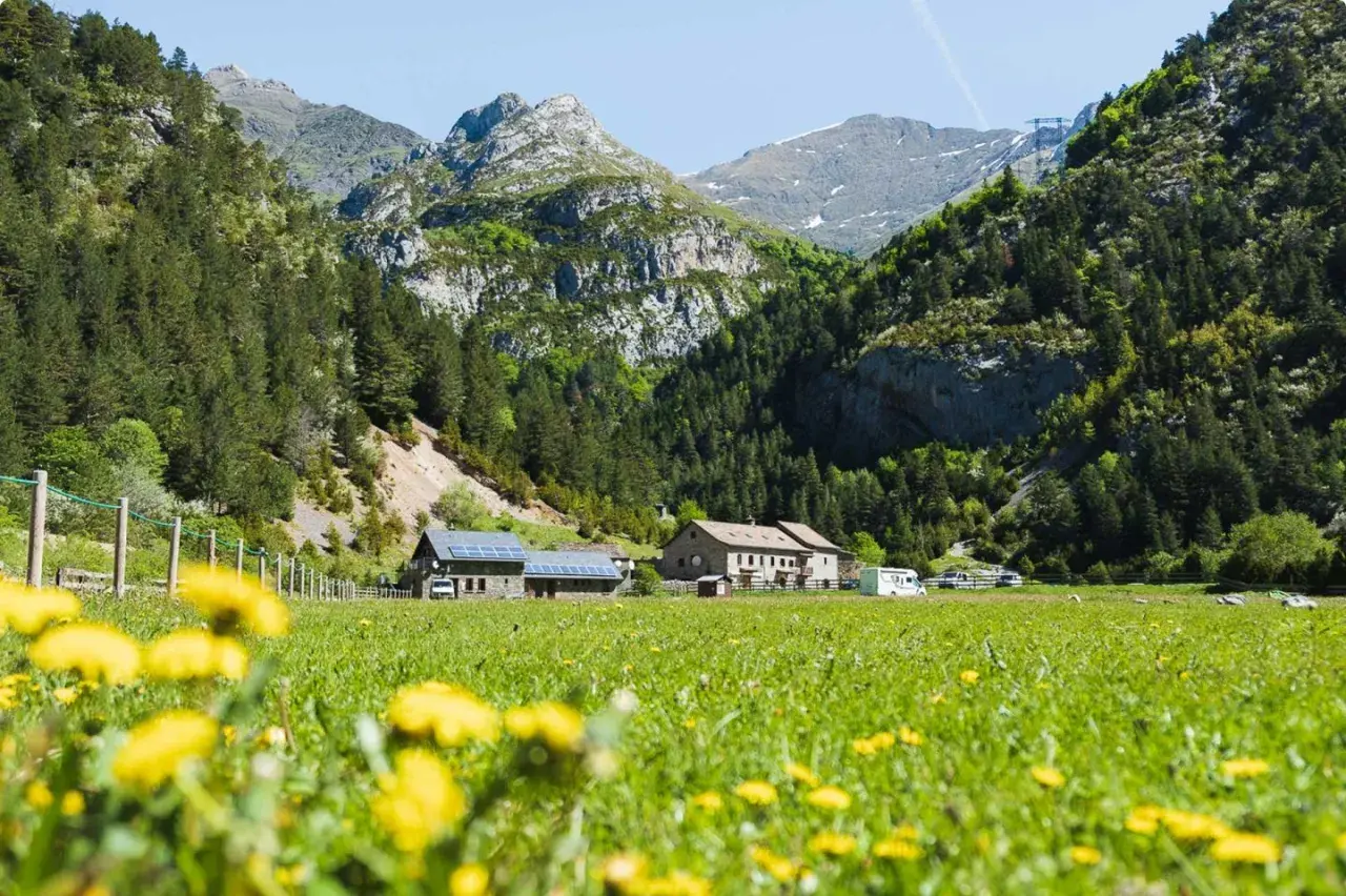 Prado florido con el Vignemale desde Bujaruelo al fondo. Casas rurales y montañas verdes bajo un cielo azul.