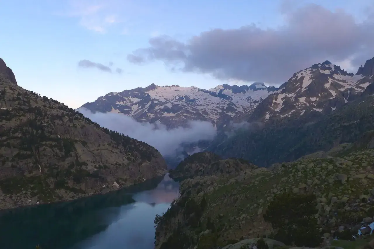 Paisaje montañoso con un lago sereno y picos nevados. Nubes bajas envuelven las montañas, creando una atmósfera mística. Un lugar idílico para explorar, como el que se encuentra en Besiberri Sud.
