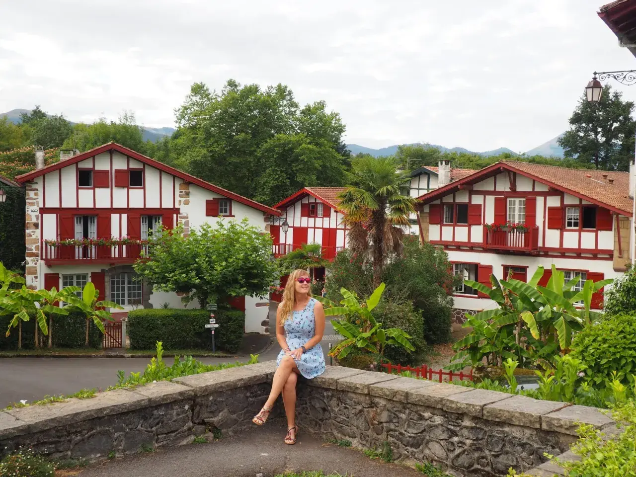 Mujer sentada en un muro de piedra, con casas blancas y rojas típicas del Pirineo Vasco al fondo.