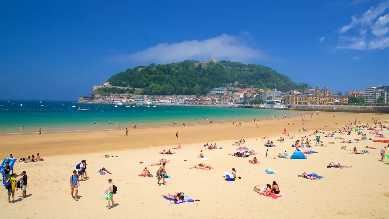 Gente disfrutando de la arena dorada y el mar turquesa en la que podría ser la mejor playa de España, con la ciudad y un monte verde de fondo.