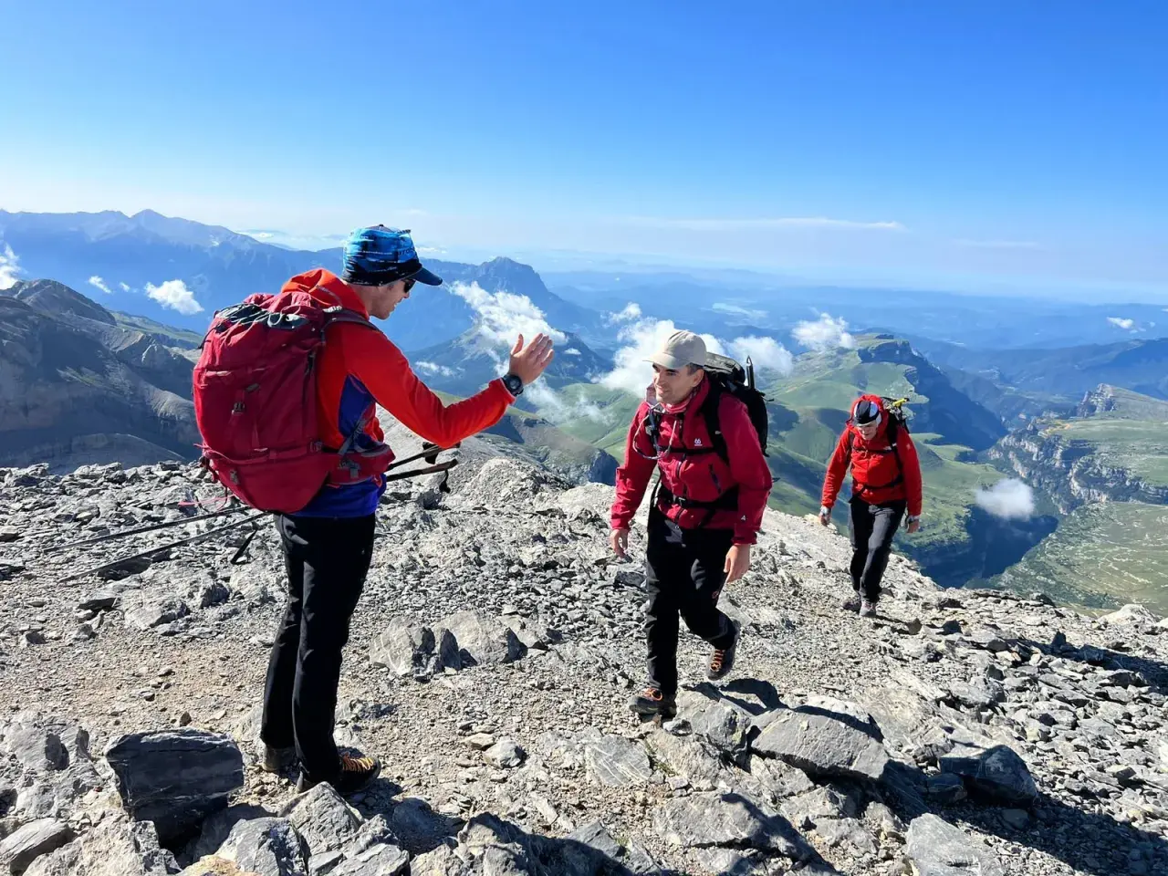 Tres excursionistas suben por una cresta rocosa en el **alt Pirineu**. El sol brilla en un cielo azul claro, con vistas a un vasto paisaje montañoso.