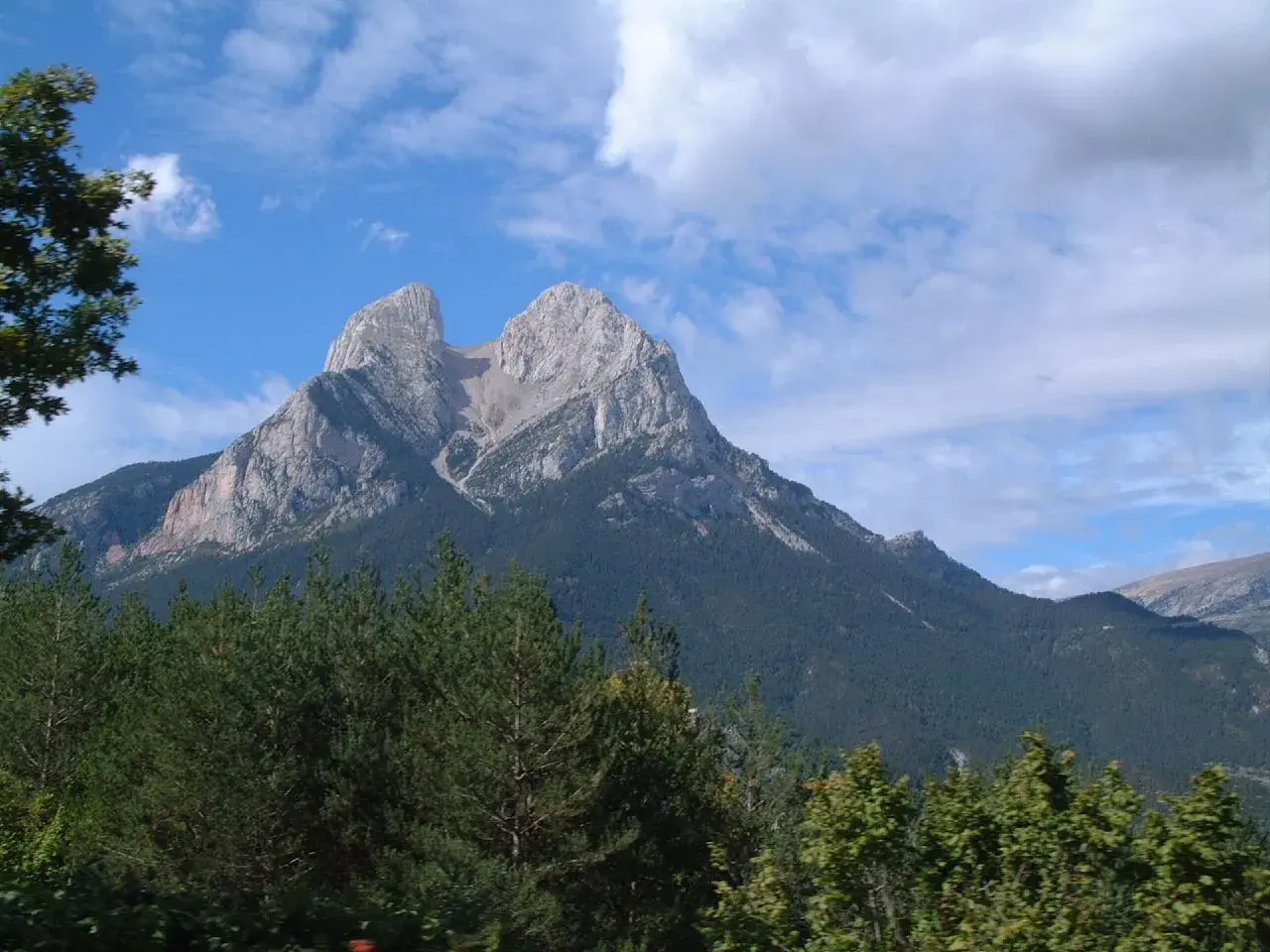 Majestuoso Pedraforca se alza imponente, su altitud desafía las nubes. Bosques verdes cubren sus faldas bajo un cielo azul.