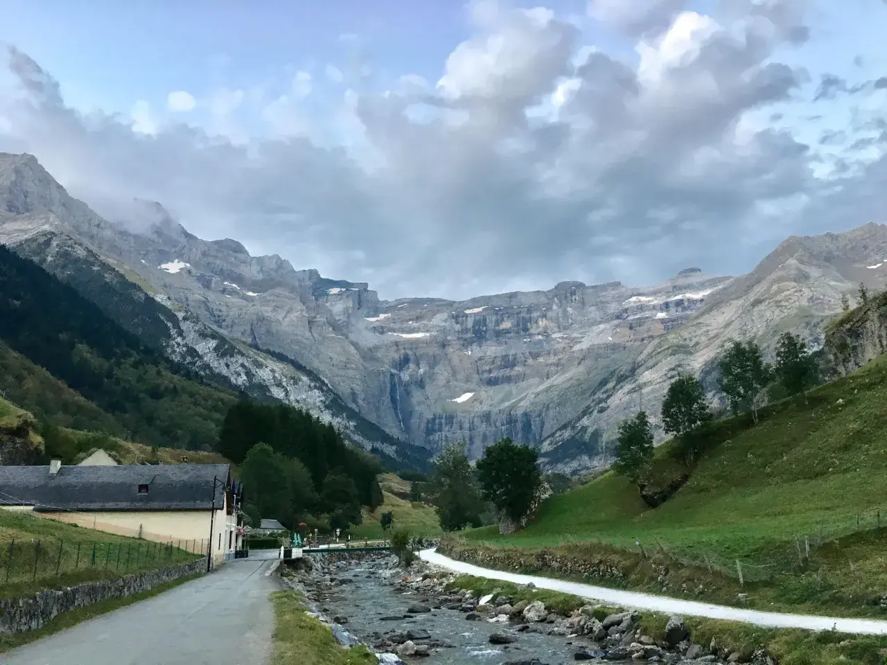 Un camino serpentea junto a un río rocoso hacia el imponente pico Taillón, con una cascada visible en la distancia y un pueblo a la izquierda.