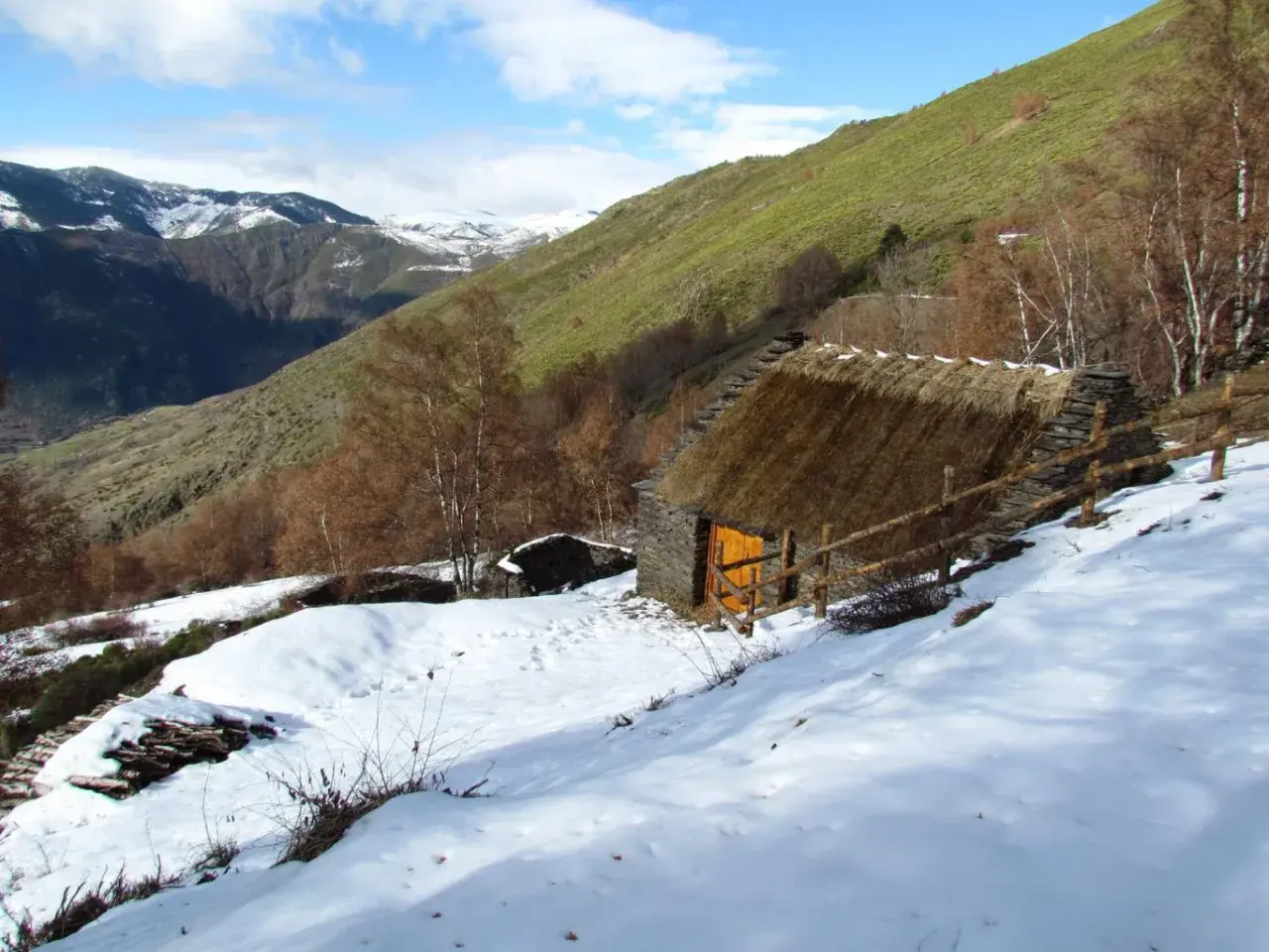 Pequeña cabaña de piedra con tejado de paja, rodeada de nieve y un paisaje montañoso. Un esterri de cardos se alza en la ladera verde.