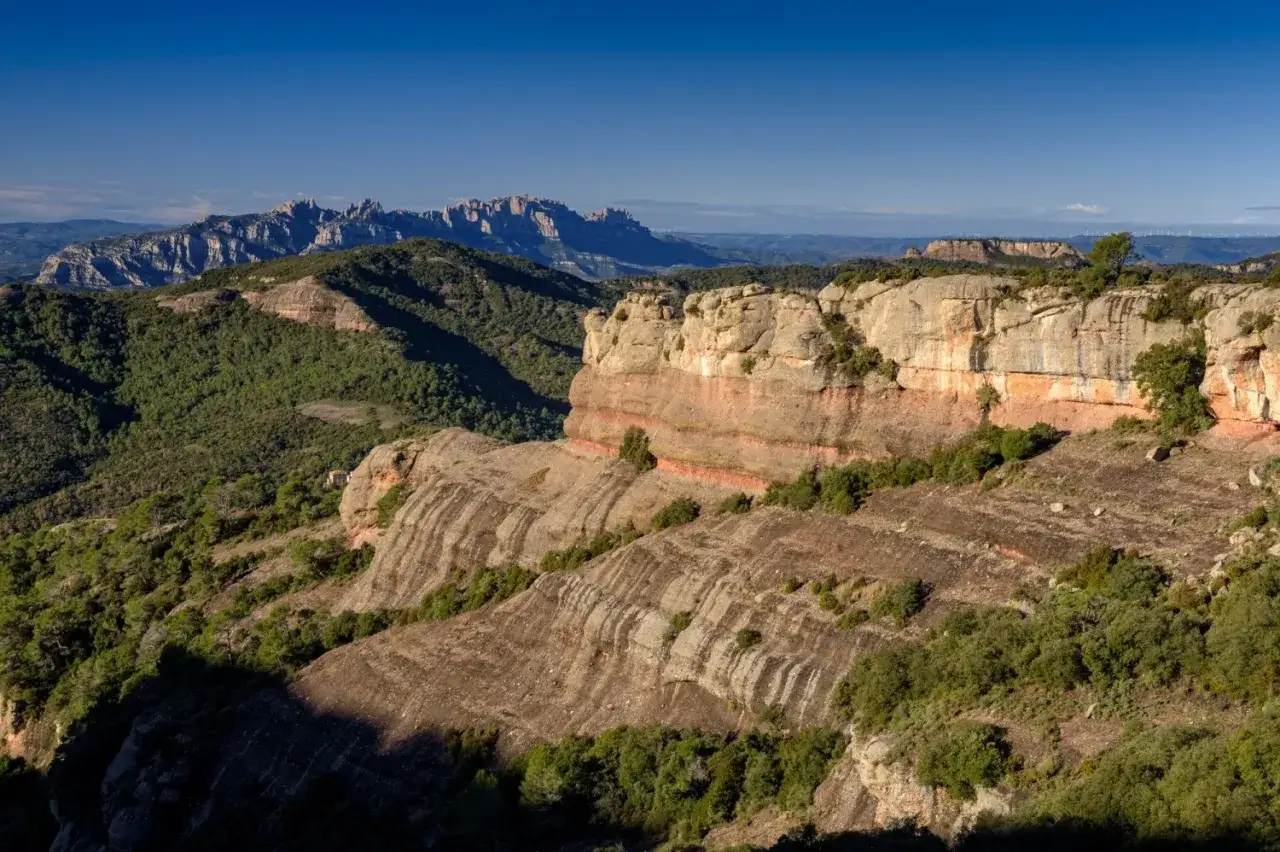 Paisaje rocoso y verde en el Montcau, con capas de roca rojiza y estriada, bajo un cielo azul intenso.