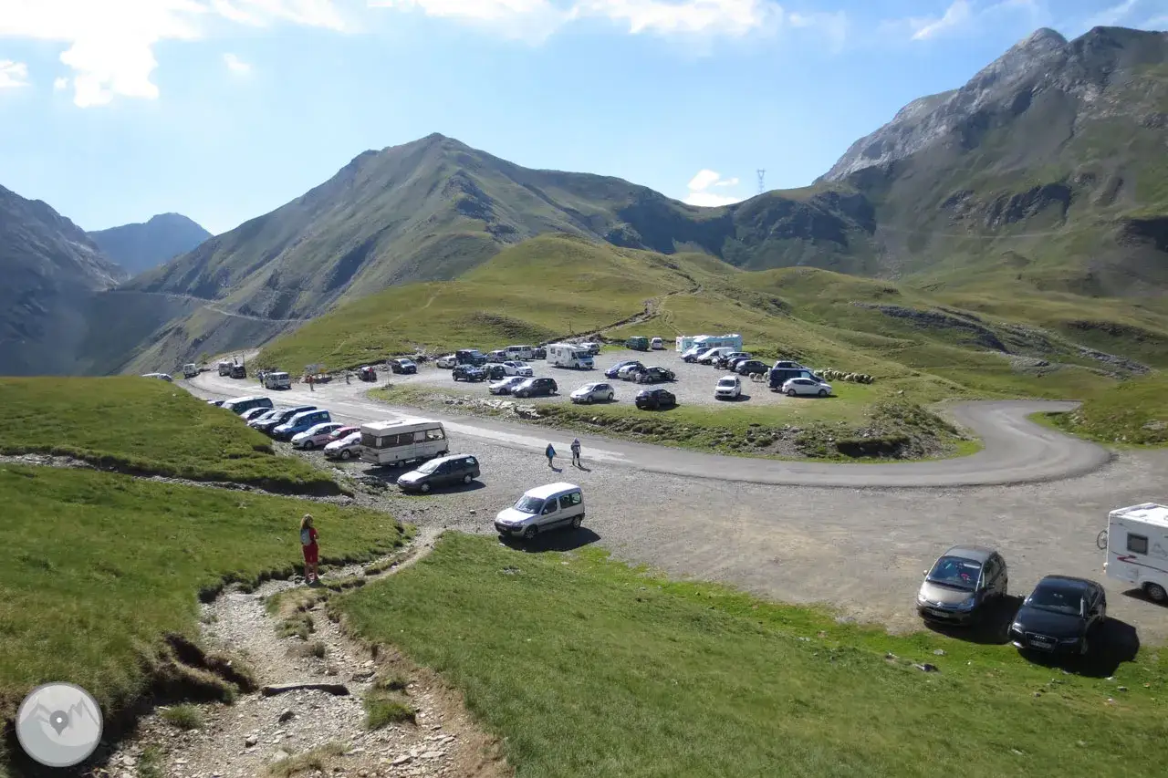 Un aparcamiento en la col de Tentes con coches y caravanas, rodeado de montañas verdes y un cielo azul.