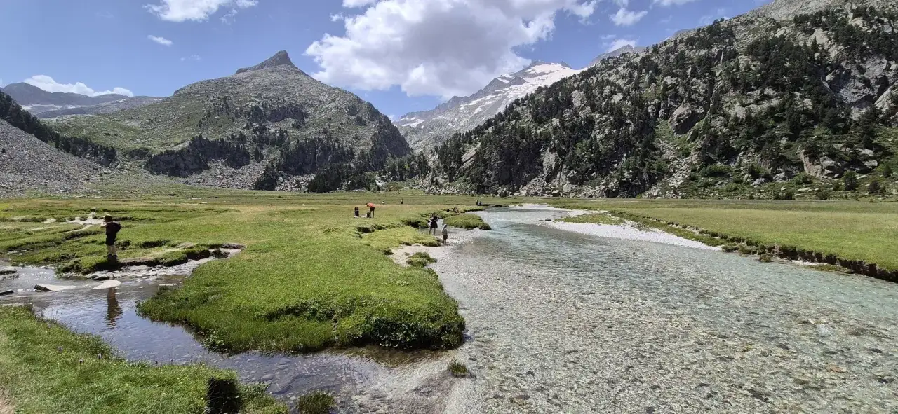 Valle verde con río cristalino y montañas imponentes, incluyendo el pico Aneto. Personas disfrutan del paisaje.