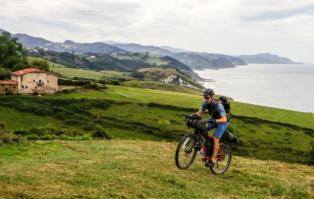 Ciclista en bikepacking carretera, pedaleando por un paisaje verde con vistas al mar y una casa rural.