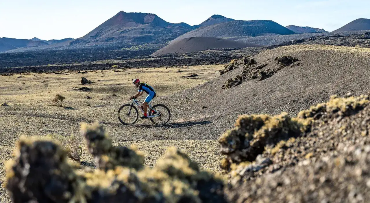 Ciclista pedaleando por un paisaje volcánico en Lanzarote en bici. El terreno árido y las montañas de fondo crean un escenario espectacular.