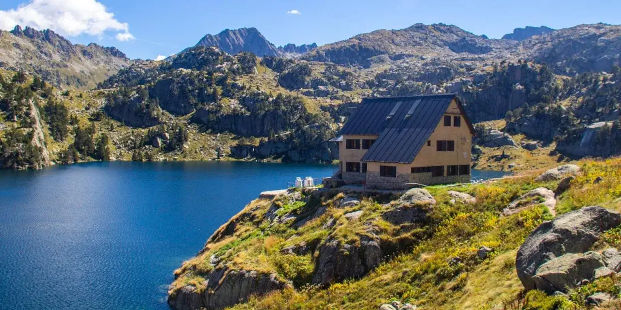 Refugio de montaña junto a un lago azul, rodeado de picos rocosos y vegetación. Ideal para la ruta de carros de foc.