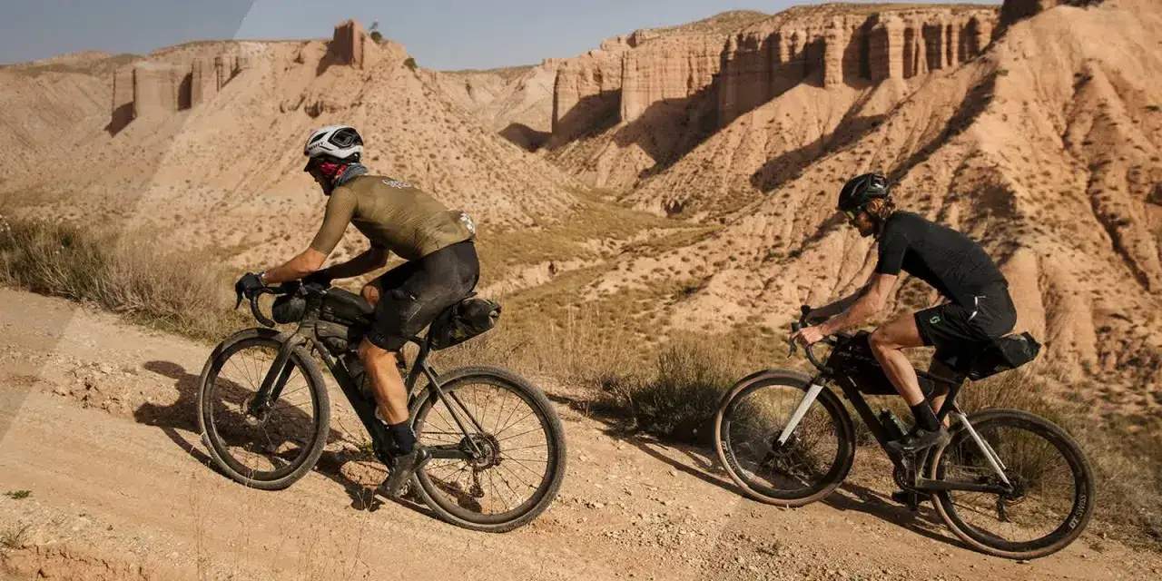 Ciclistas pedalean por un camino de tierra en un paisaje desértico de badlands. El terreno de badlands gravel presenta formaciones rocosas erosionadas y vegetación escasa.