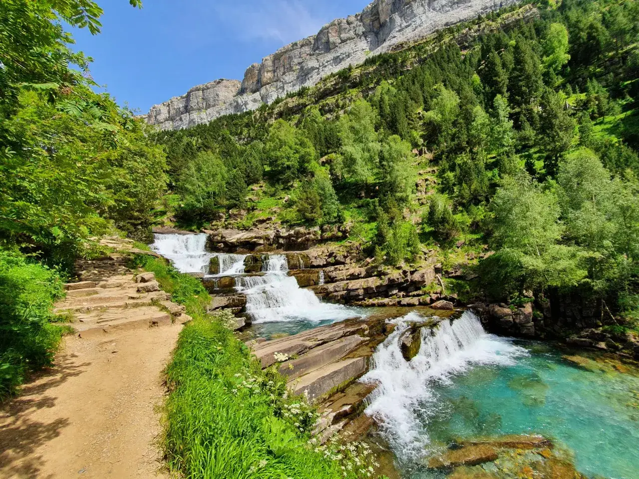 Sendero de grava junto a las gradas de soaso, cascadas de agua turquesa cayendo entre rocas, rodeadas de vegetación exuberante y un imponente acantilado.