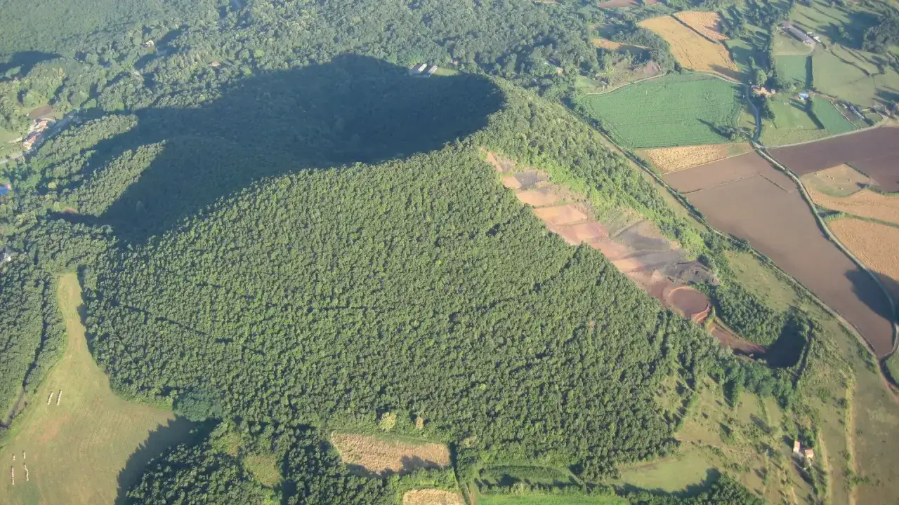 Vista aérea del volca de la Crosa, un cono volcánico cubierto de vegetación exuberante, con campos agrícolas y un pueblo en el paisaje circundante.