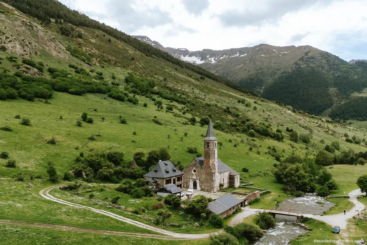Iglesia románica en el Val d'Aran, rodeada de verdes prados y montañas. Un río serpentea cerca.