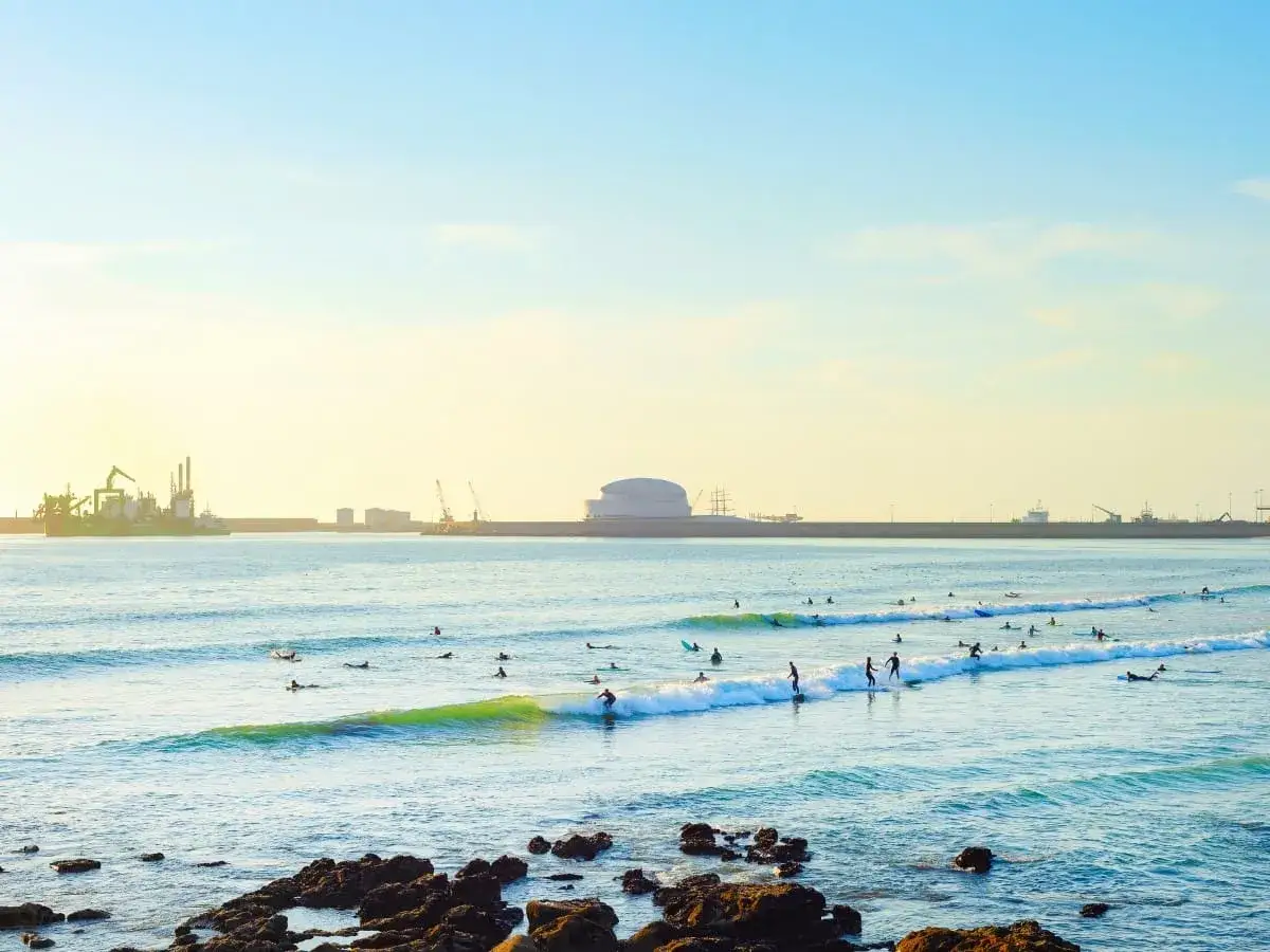 Ondas perfectas para surfear en la playa Oporto Matosinhos. El sol ilumina el mar y el puerto industrial al fondo.