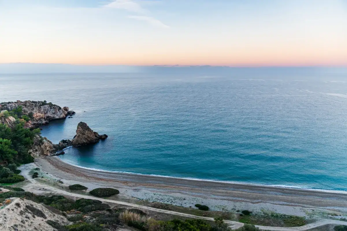Vista aérea de una cala rocosa y una playa de guijarros en las playas de Málaga al amanecer. El mar azul turquesa se encuentra con el cielo pastel.