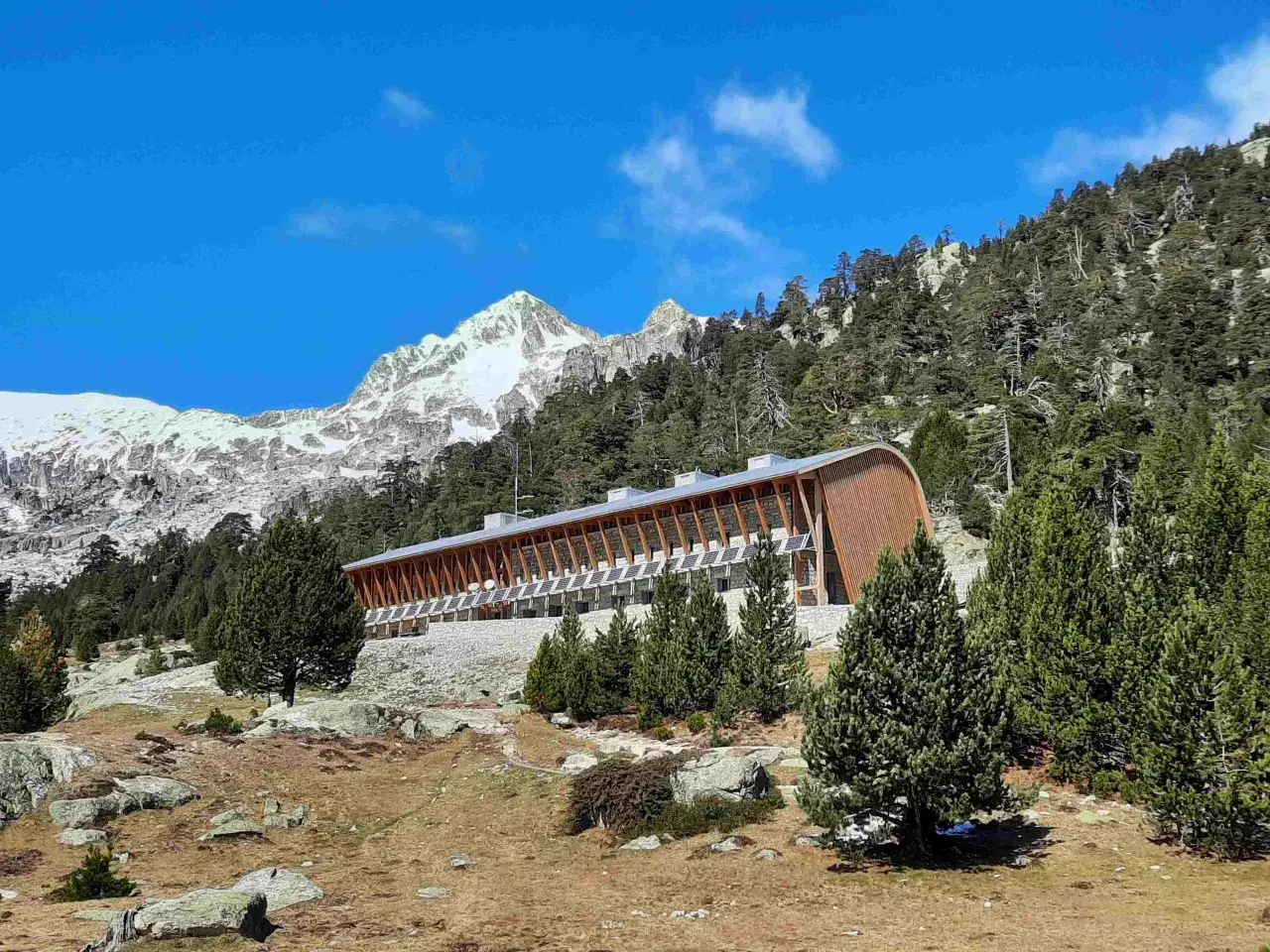 Edificio moderno de madera y piedra en un paisaje montañoso, con picos nevados al fondo. El refugio Vignemale se integra en la naturaleza.