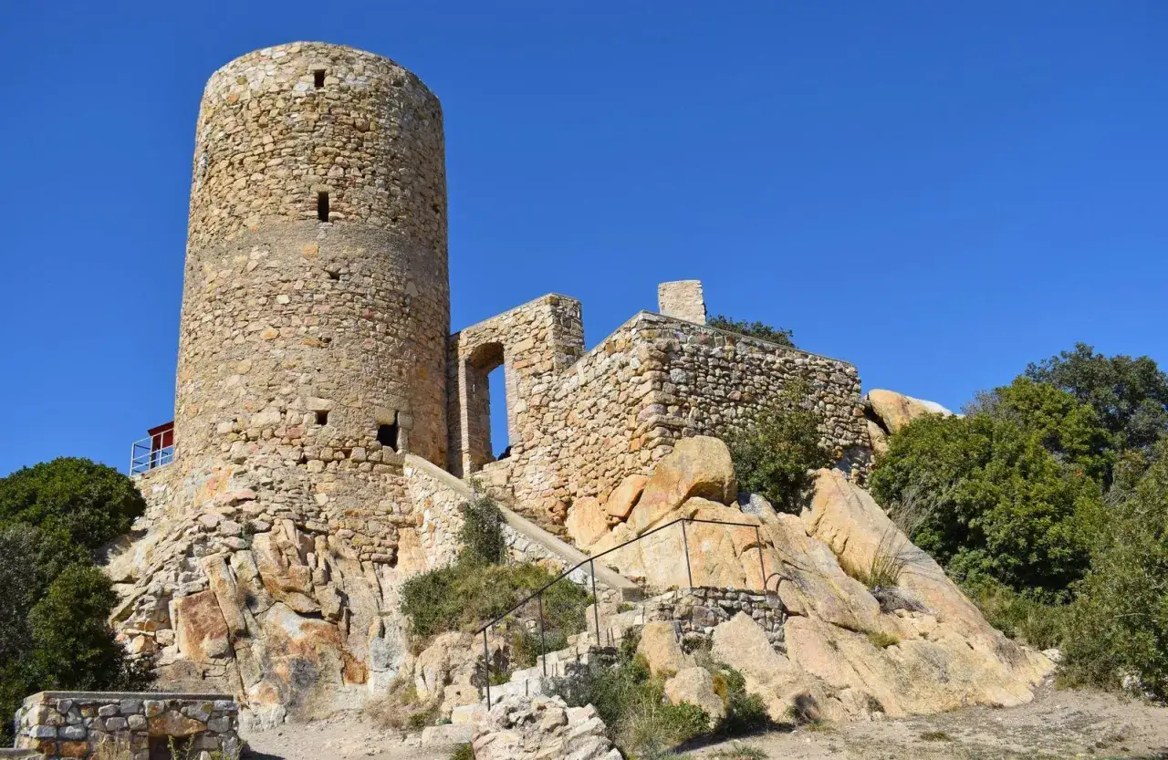 El castillo de Burriac, una torre circular de piedra y ruinas, se alza sobre una colina rocosa bajo un cielo azul intenso.