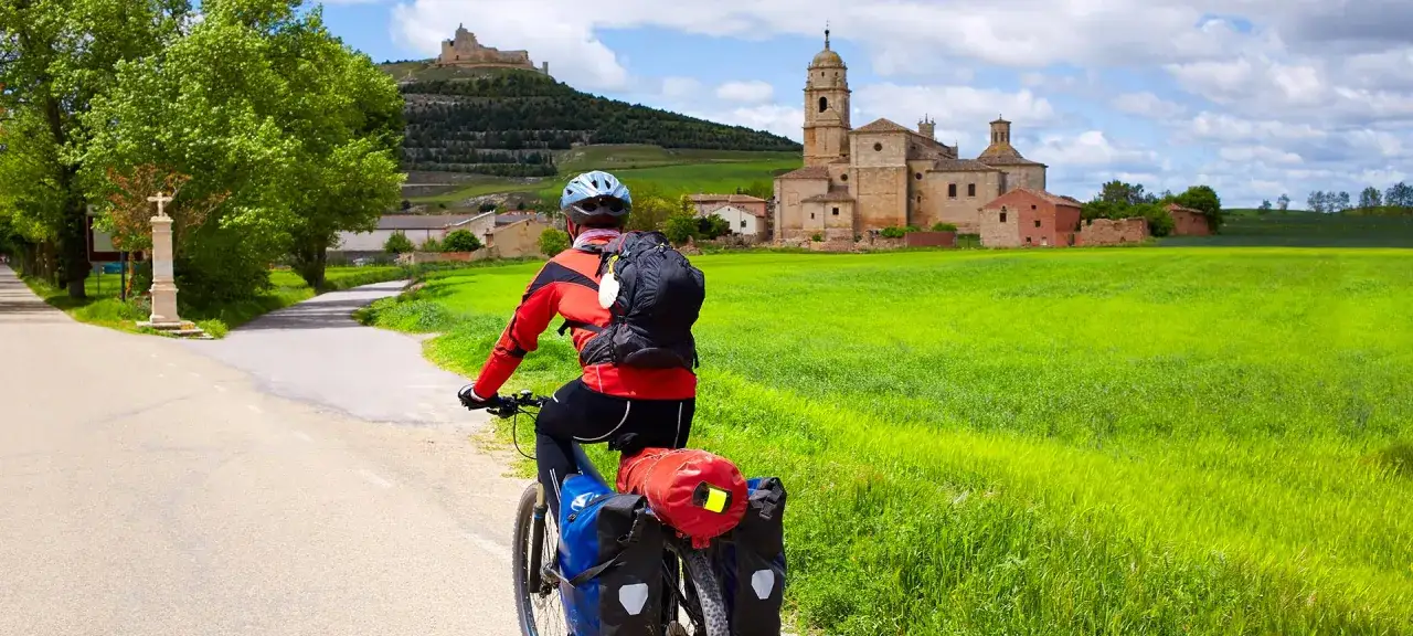 Ciclista con alforjas, listo para su bicicleta para Camino de Santiago, pasa junto a un campo verde y una iglesia antigua.