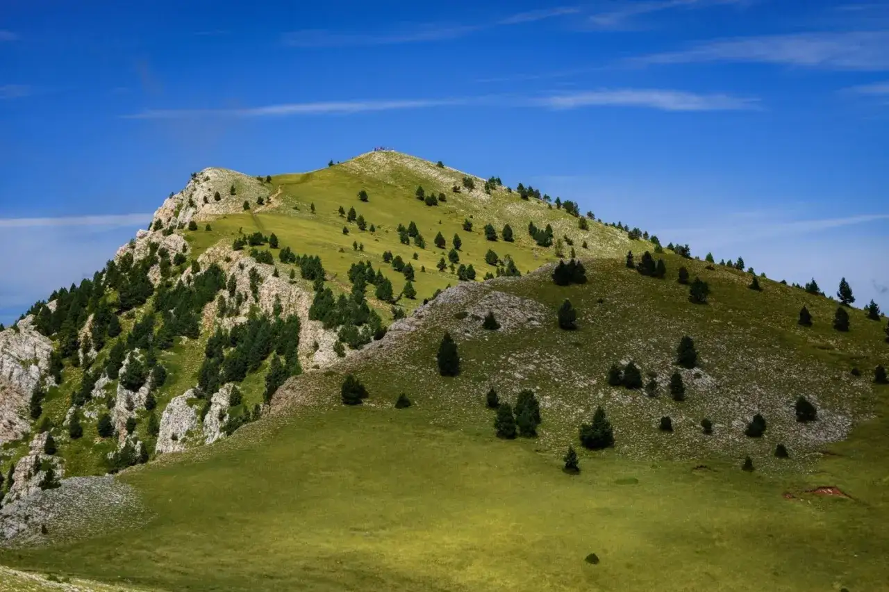 Majestuosa Serra d'Ensija bajo un cielo azul intenso, con laderas verdes salpicadas de pinos y rocas.