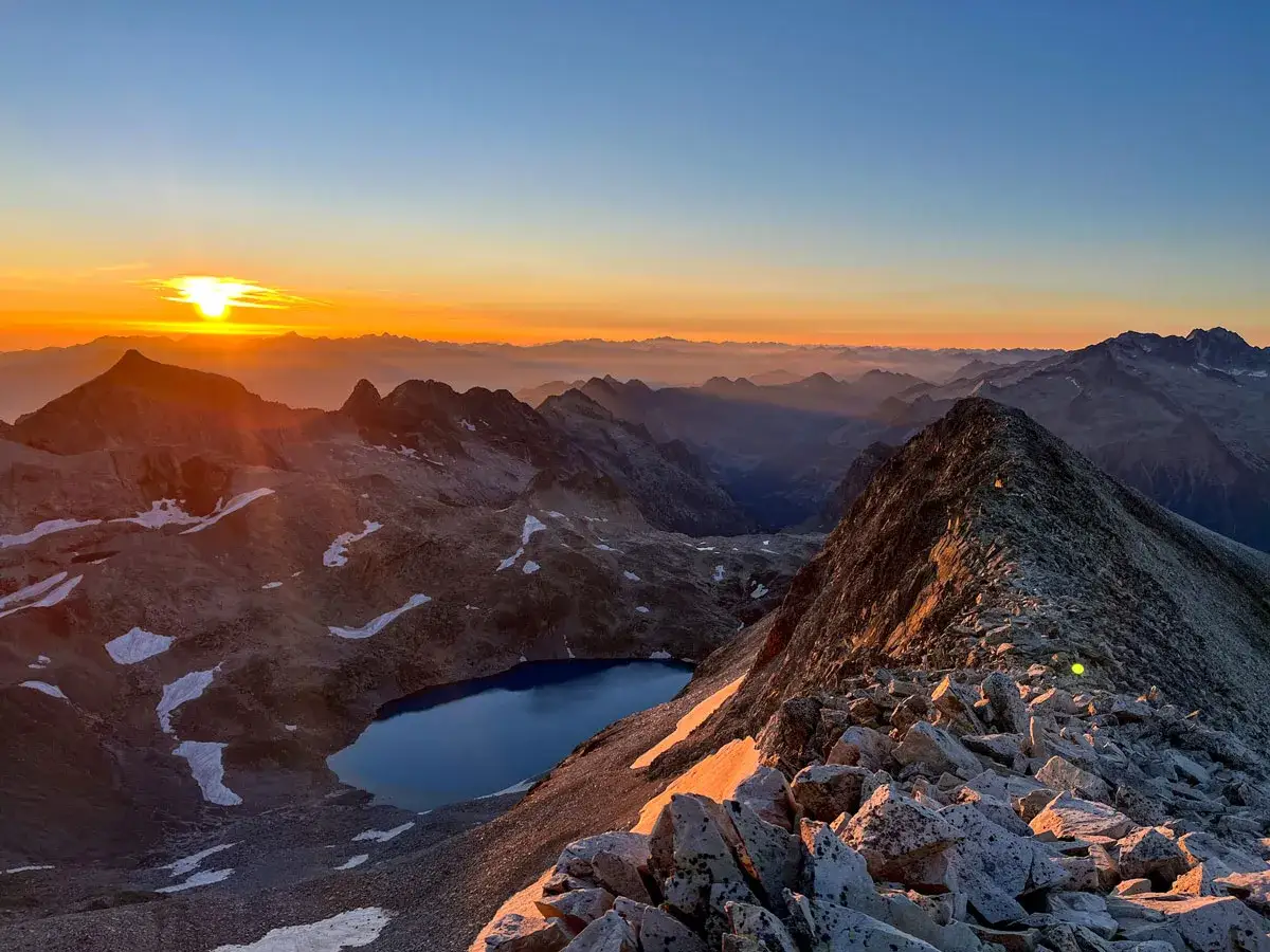 Amanecer dorado sobre un lago azul alpino, con el sol asomando tras el pico perdiguero y montañas en capas.