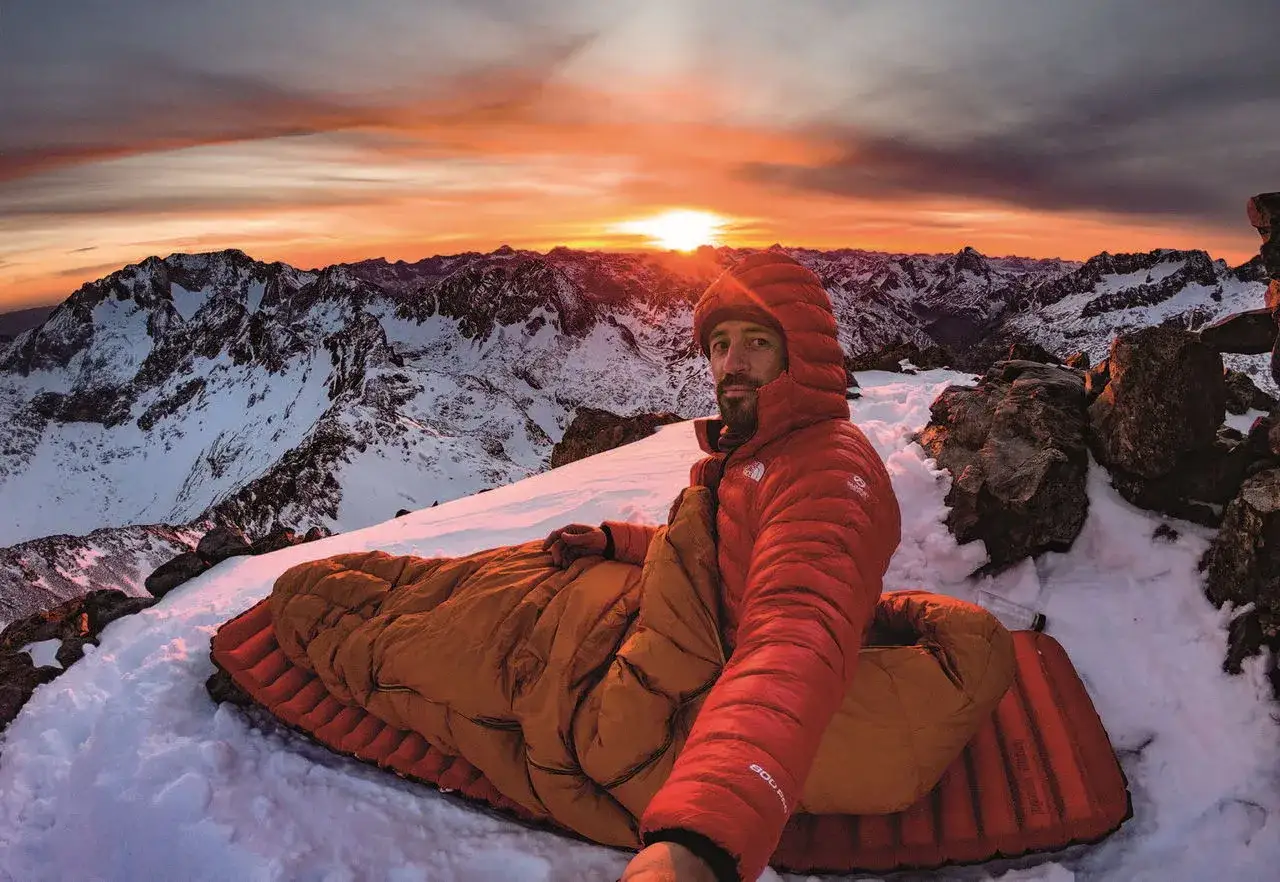 Hombre en saco de dormir en la cima de una montaña nevada, disfrutando de un amanecer espectacular. Una experiencia de acampada libre Francia inolvidable.