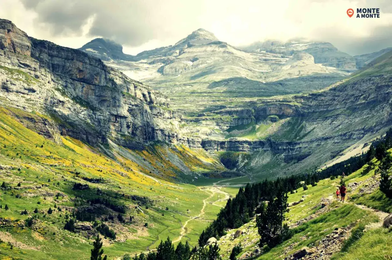 Un valle verde y florido se abre paso entre imponentes acantilados, revelando la majestuosidad de la brecha de Rolando. Dos excursionistas recorren un sendero serpenteante.