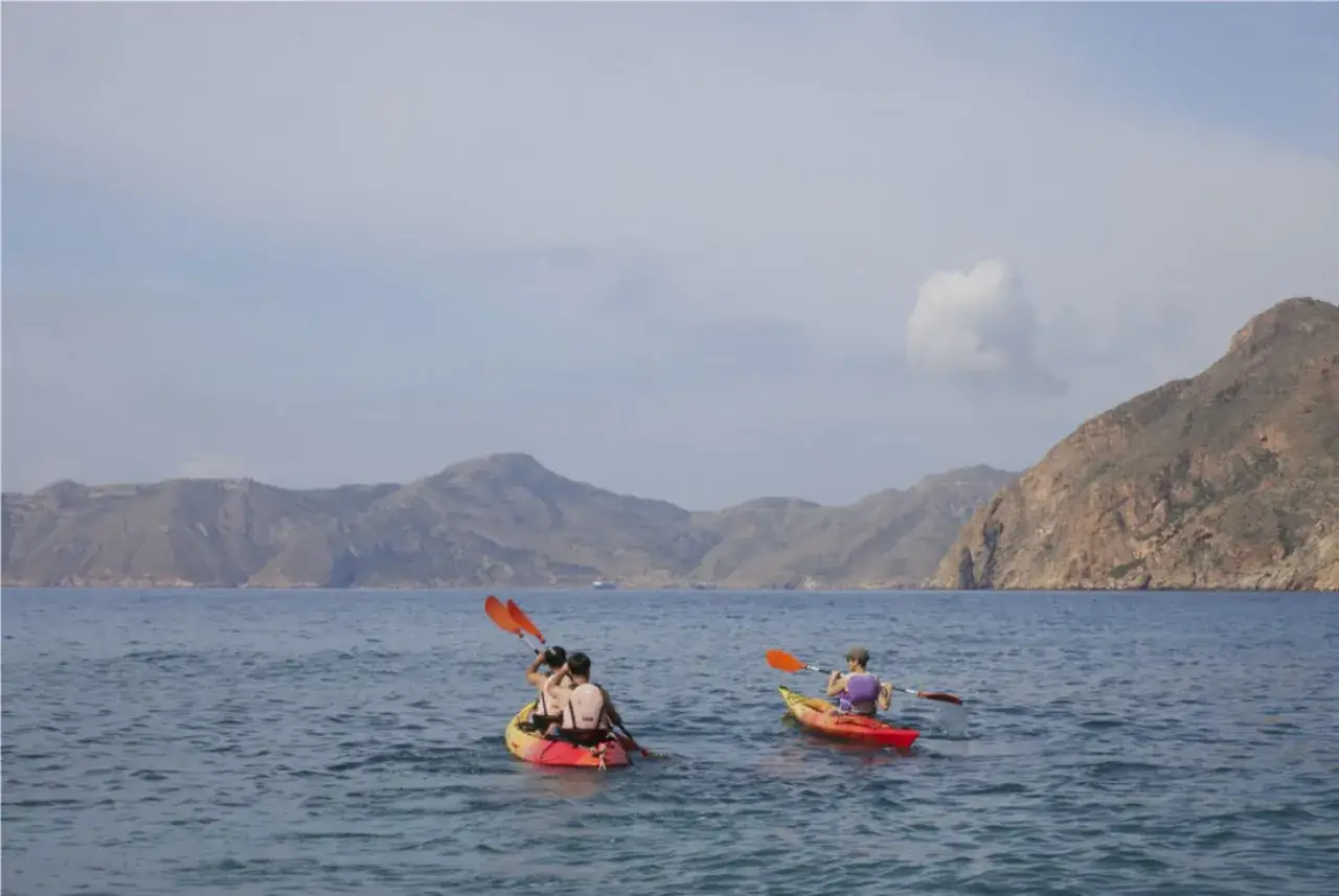 Personas en kayaks disfrutan de un día soleado en el mar, con montañas de fondo. Una aventura de camping en Andalucía.