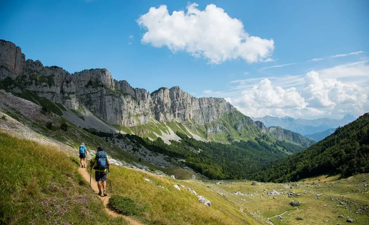 Dos excursionistas caminan por un sendero en el Pirineo Vasco, rodeados de imponentes acantilados y exuberante vegetación.