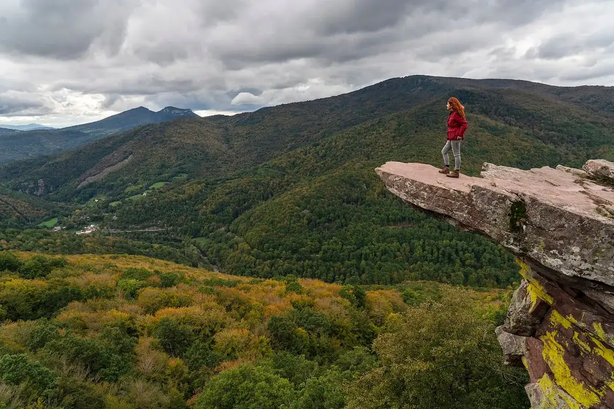 Mujer con cabello rojizo contempla el paisaje otoñal desde un saliente rocoso en el Pirineo Navarro.