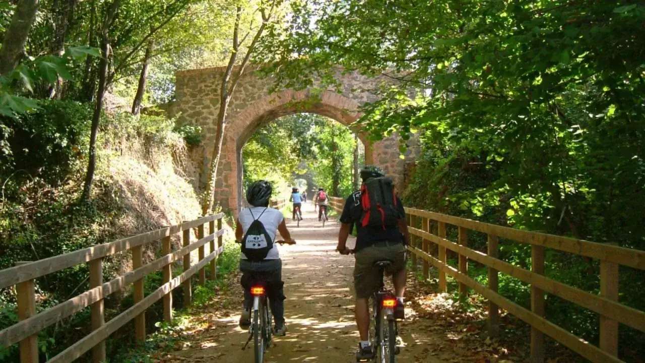 Ciclistas disfrutan de la vía verde Olot Girona, pedaleando bajo un puente de piedra rodeado de frondosa vegetación.