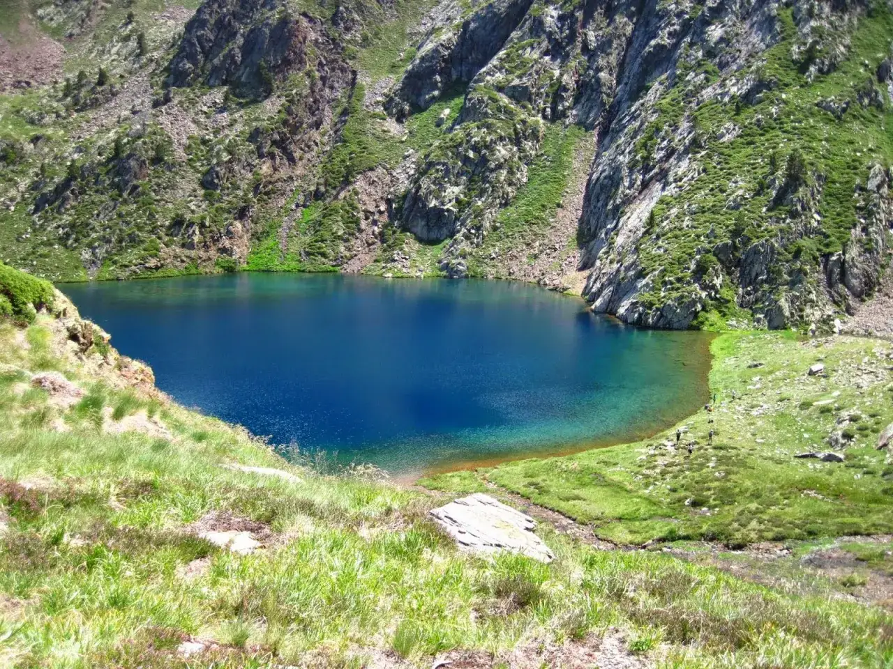 Lago azul intenso en el alt pirineu, rodeado de montañas rocosas y vegetación verde.