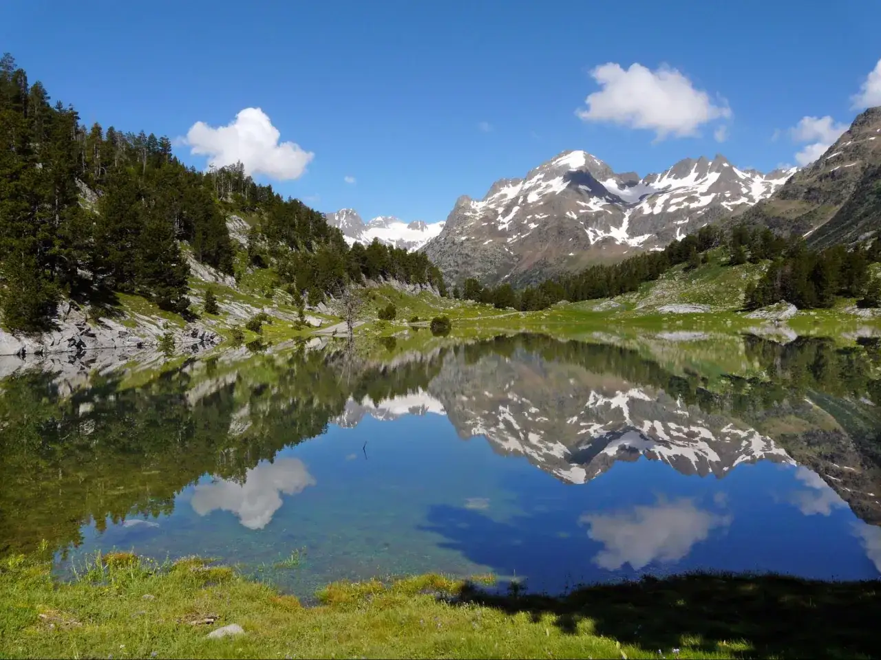 Espejo de agua en el Pirineo Aragonés, reflejando montañas nevadas y un cielo azul con nubes.
