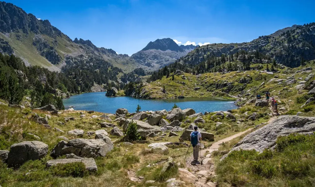 Senderistas disfrutan de un día soleado en el Valle de Arán, rodeados de montañas, un lago azul y rocas.