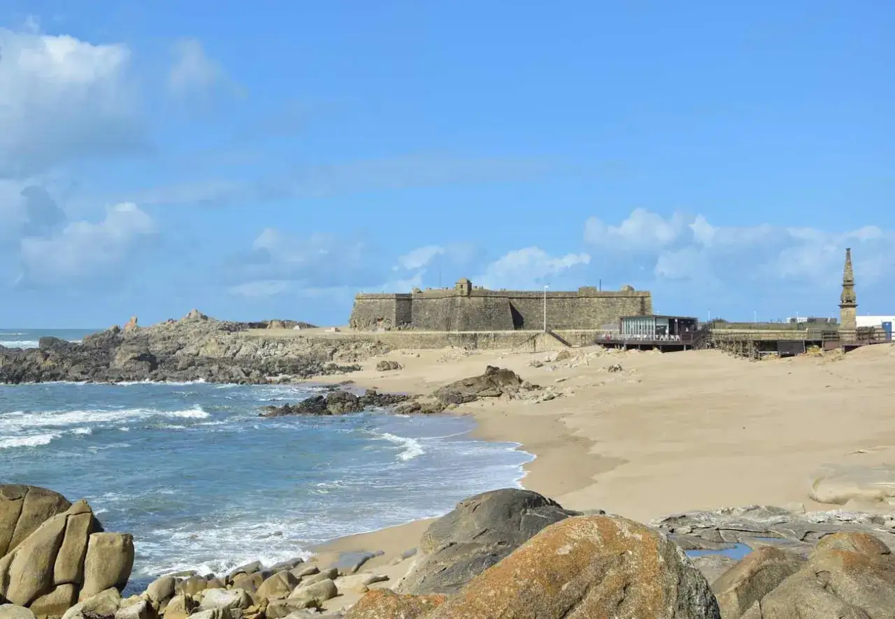 Fortaleza histórica junto a las playas de Oporto, con rocas y mar azul bajo un cielo nublado.