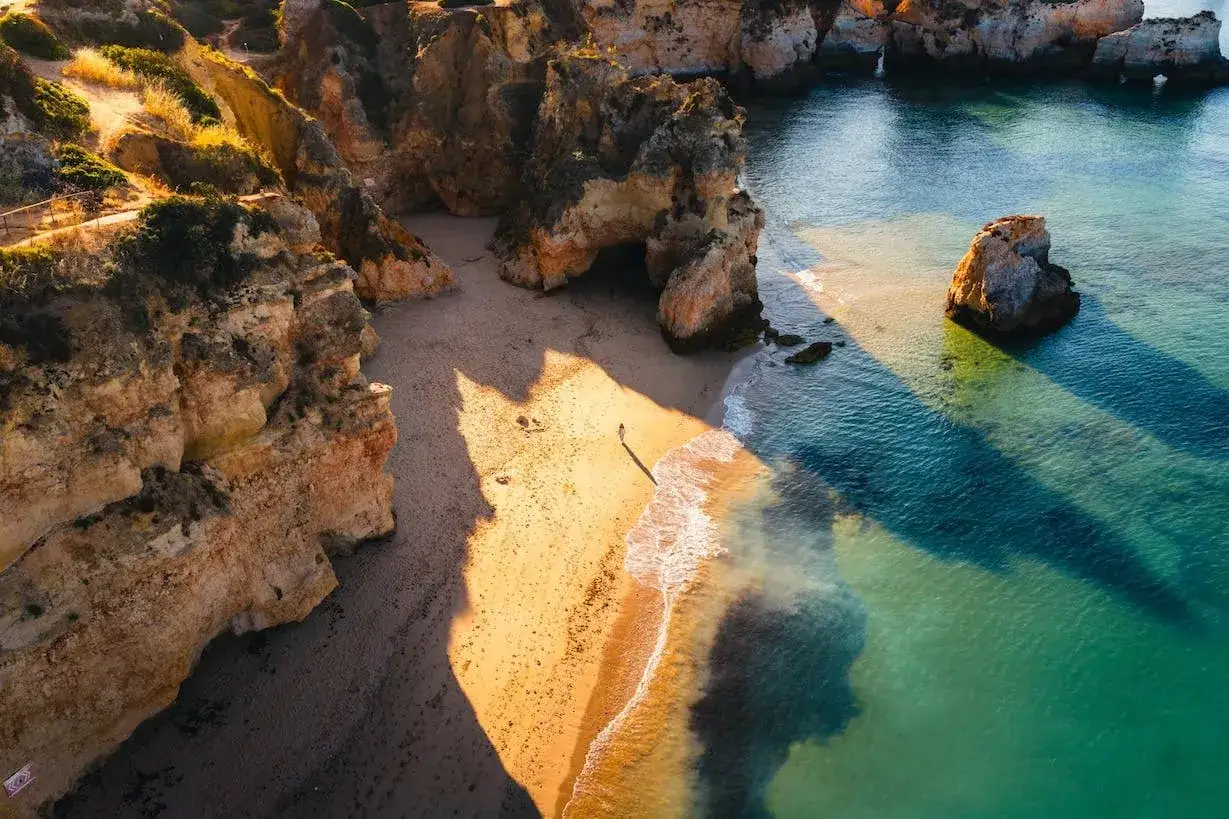 Una persona camina por una playa dorada en las calas del Algarve, con acantilados ocres y aguas turquesas bañadas por el sol.