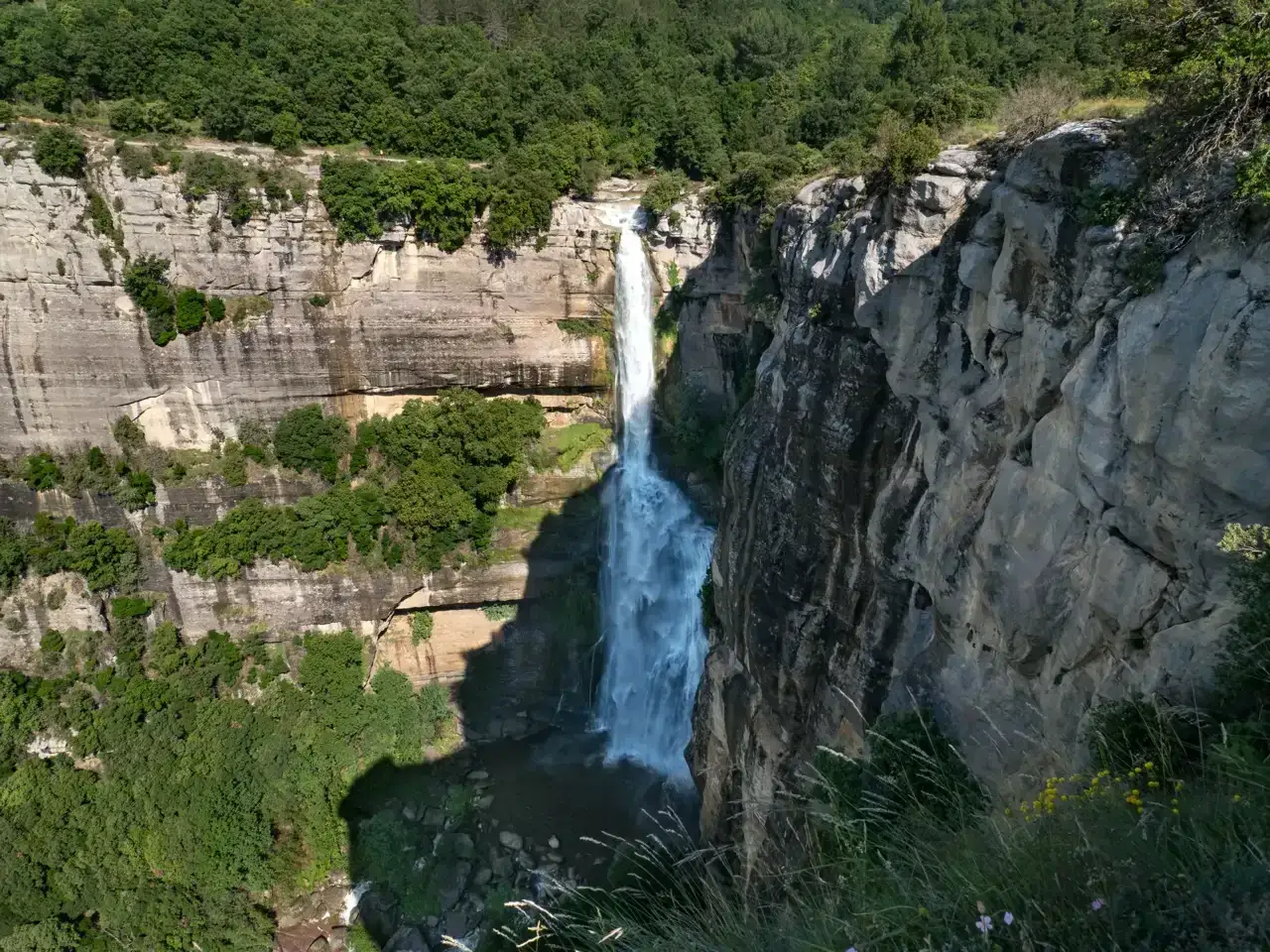 Impresionante cascada cayendo por un acantilado rocoso, parte de la ruta Rupit. Vegetación exuberante rodea la caída de agua.