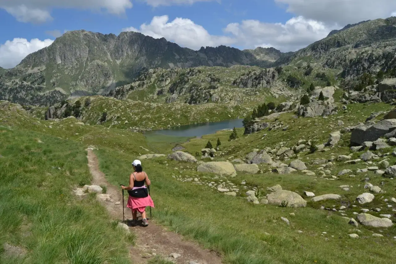 Mujer camina por senderos de montaña, disfrutando de las rutas vielha y el paisaje alpino con un lago azul.